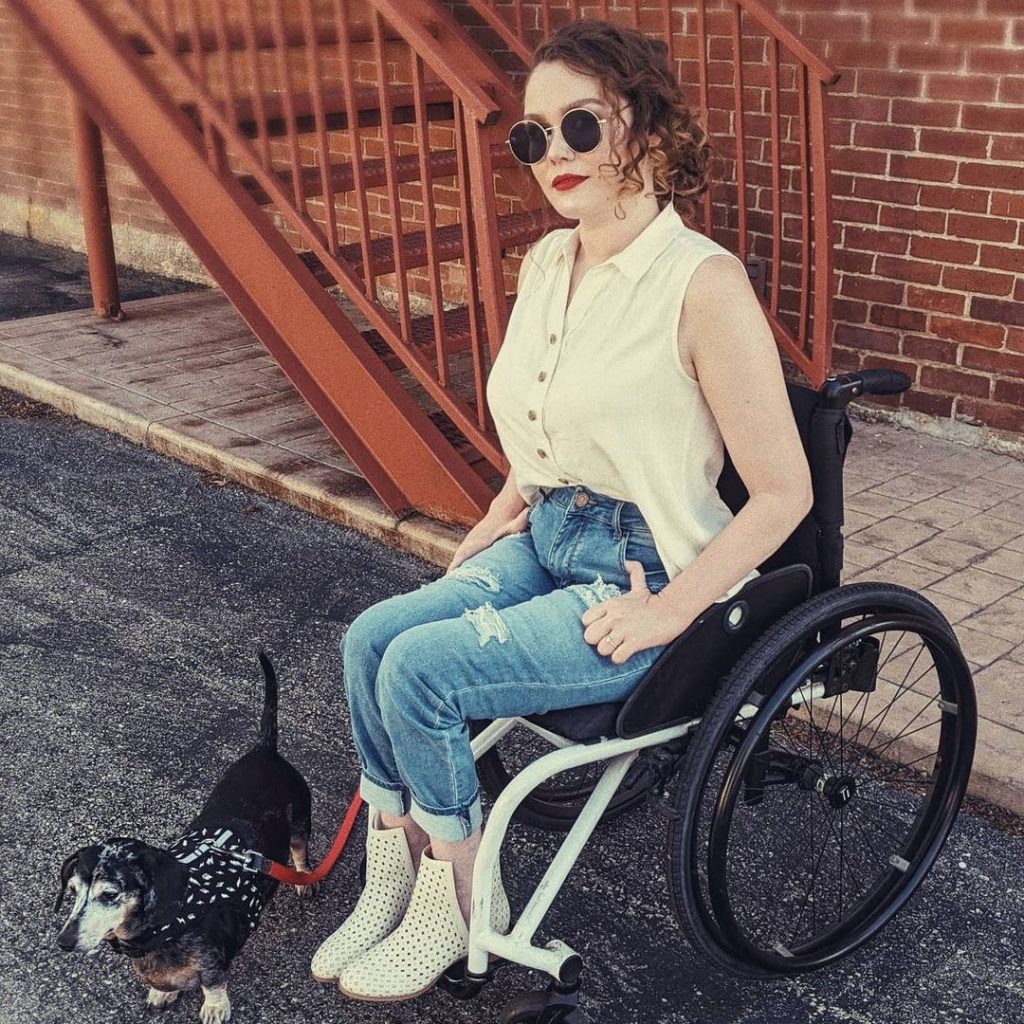 Monica Engle Thomas, a white woman with curly auburn hair, is shown in this photo wearing a white Yarrow sleeveless button down that Tracy designed. Monica sits in her black and white manual wheelchair. She also wears sunglasses and jeans, while holding the leash to her small dog. 