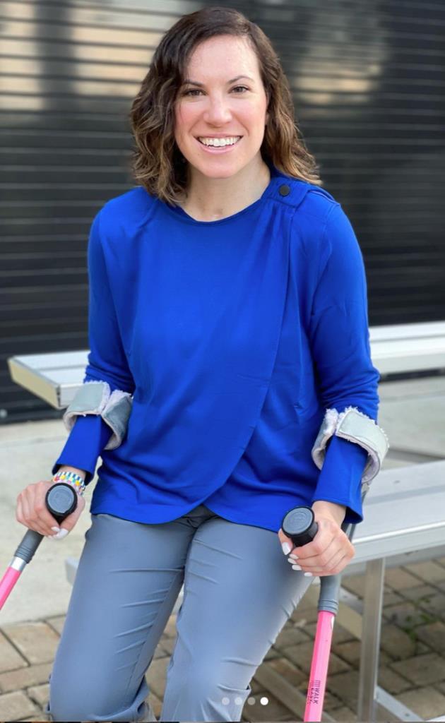 Molly Farrell, a white woman with brown hair, is shown in this photo wearing ULEX, one of the brands Tracy designed and helped launch. Molly is wearing a royal blue wrap cardigan and gray pants, while seated on bleachers. She is smiling brightly and her pink forearm crutches are visible in the photo. 